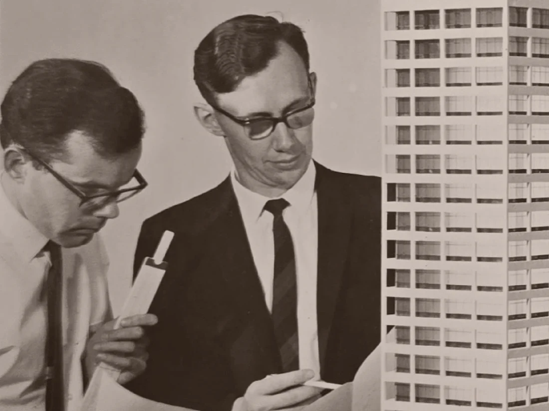 Archive photograph of two designers in suits examining a scale model of an Auckland apartment tower